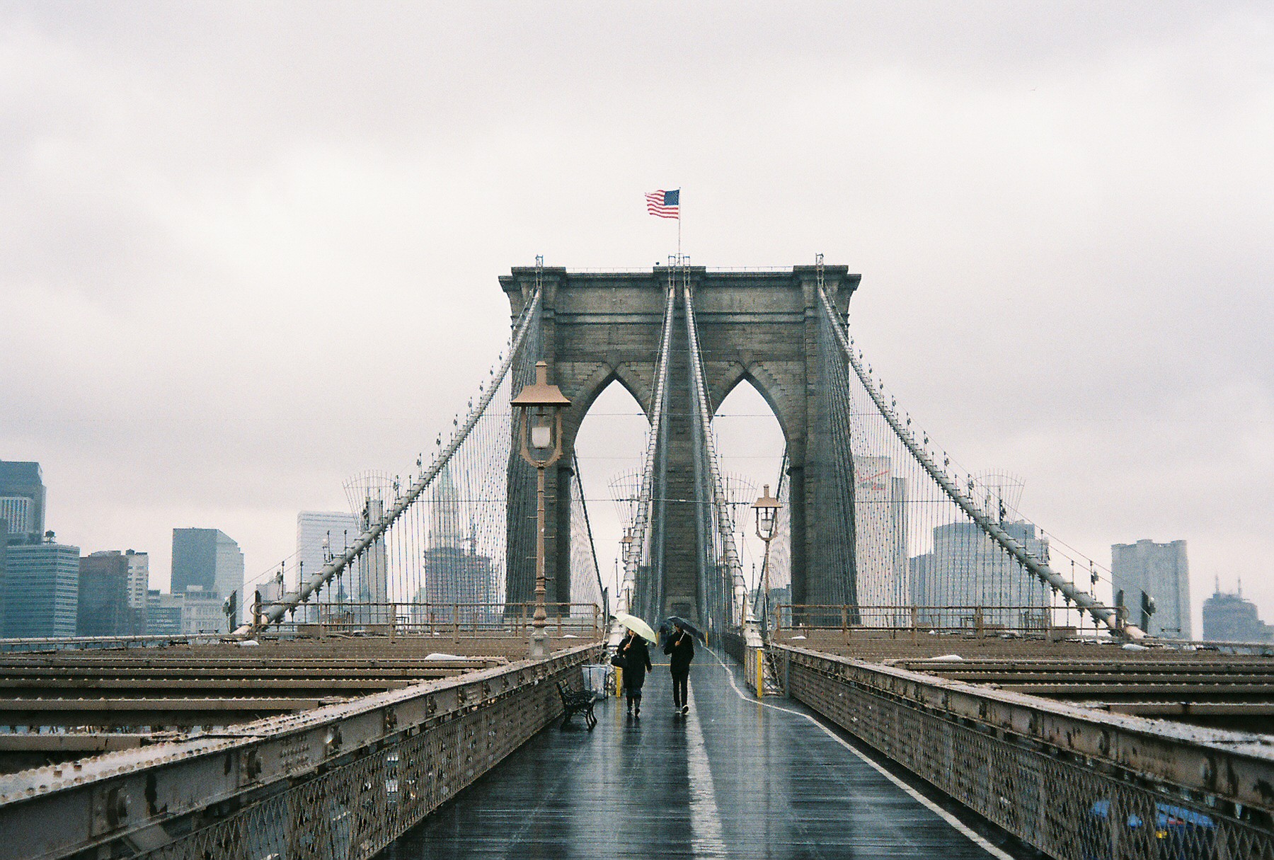 A photo of two people walking on the Brooklyn Bridge in New York City on a rainy day, with an American flag waving in the wind at the top.
