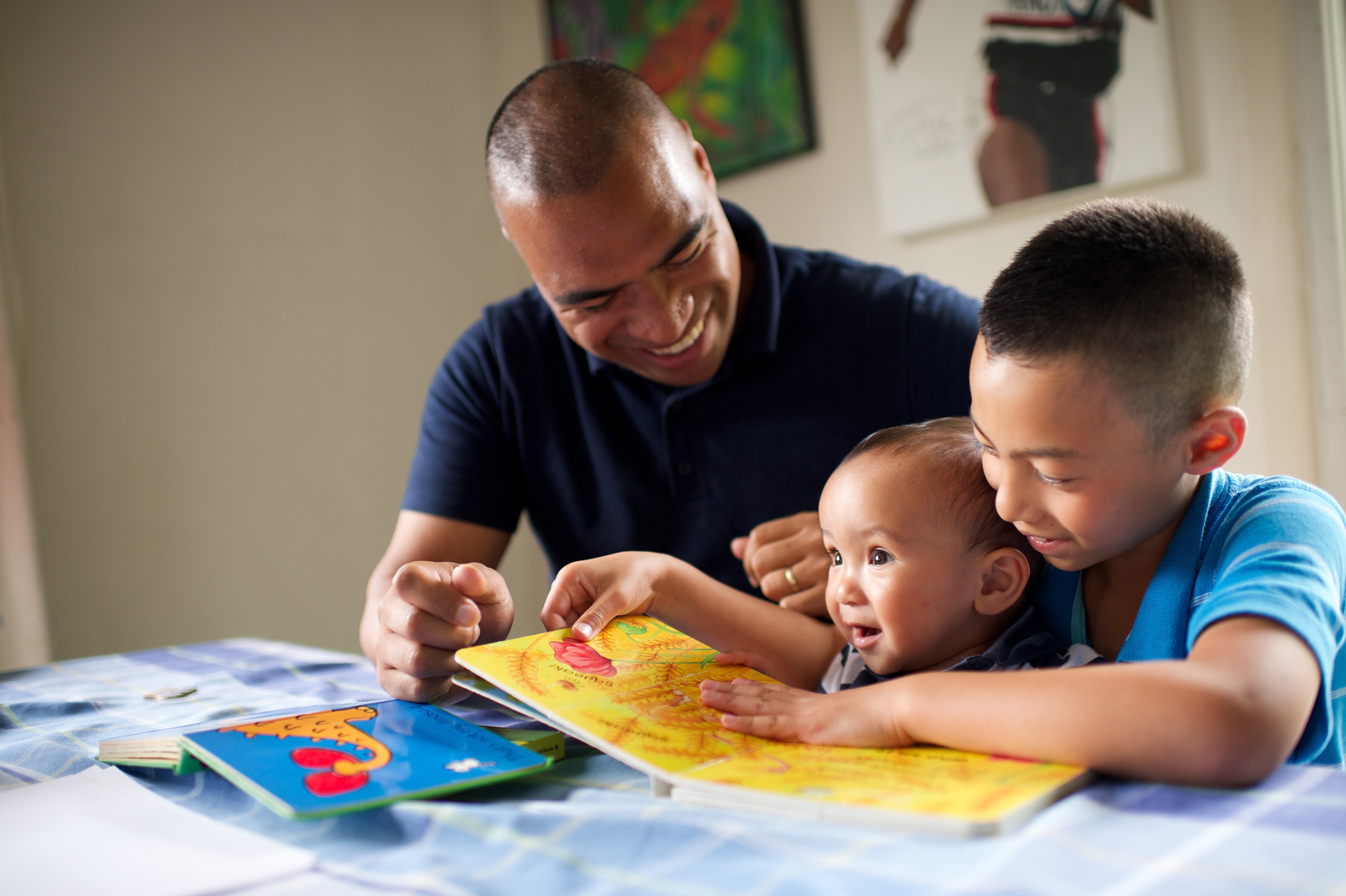 A father sits at a table with his two sons and reads a picture book to them.