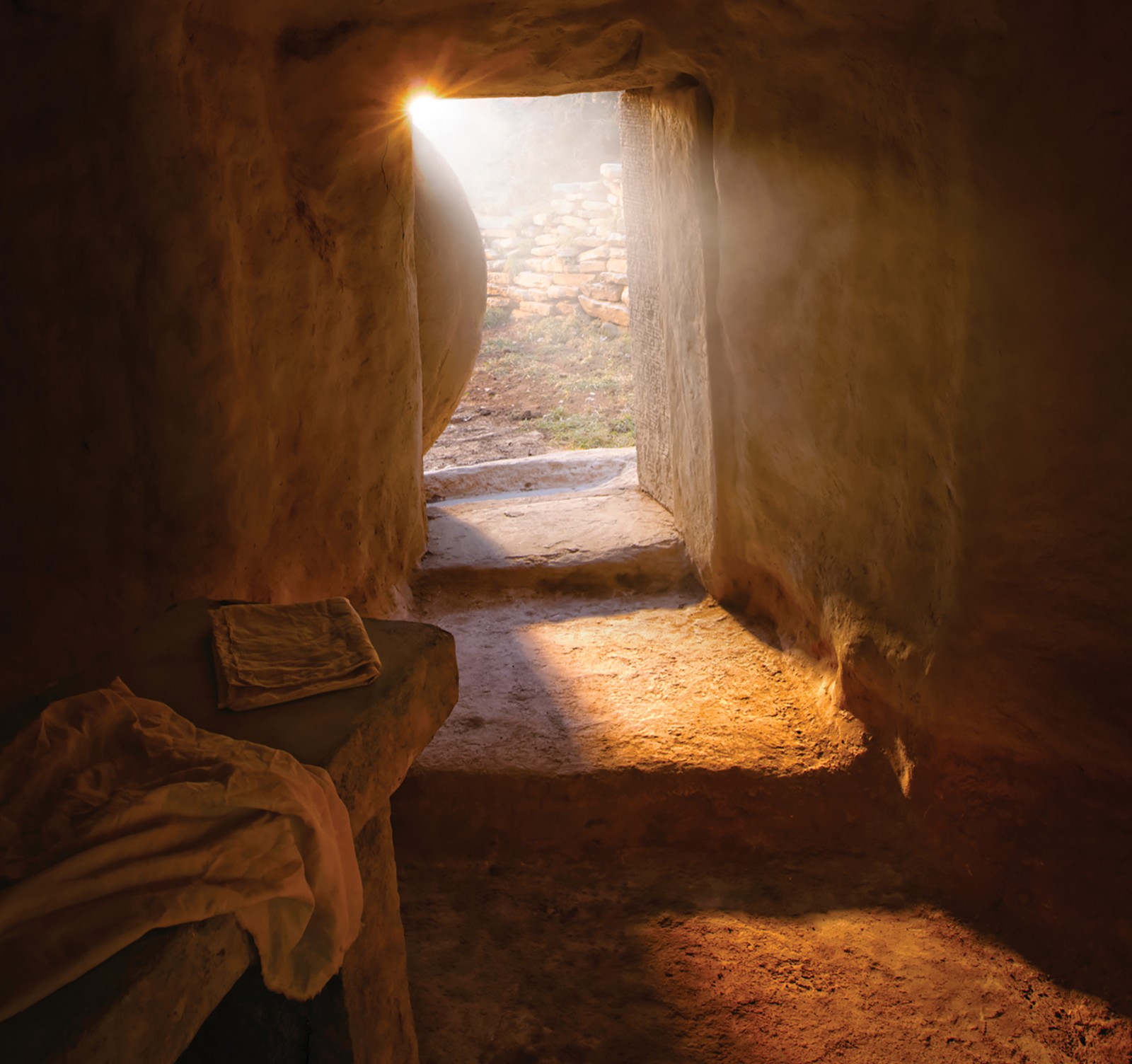 A tomb with stone steps leading to an empty burial bed and discarded wrappings. At the entrance the stone has been rolled away to reveal rays of sunlight and a partial view of outside the tomb.