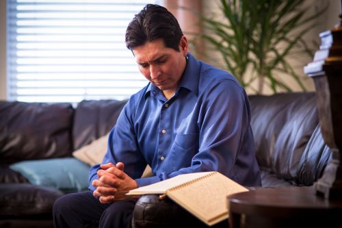 A man clasps his hands and rests them on his knee while he sits in prayer with his journal open.