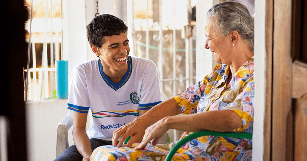 Um rapaz se senta com uma mulher idosa e riem juntos