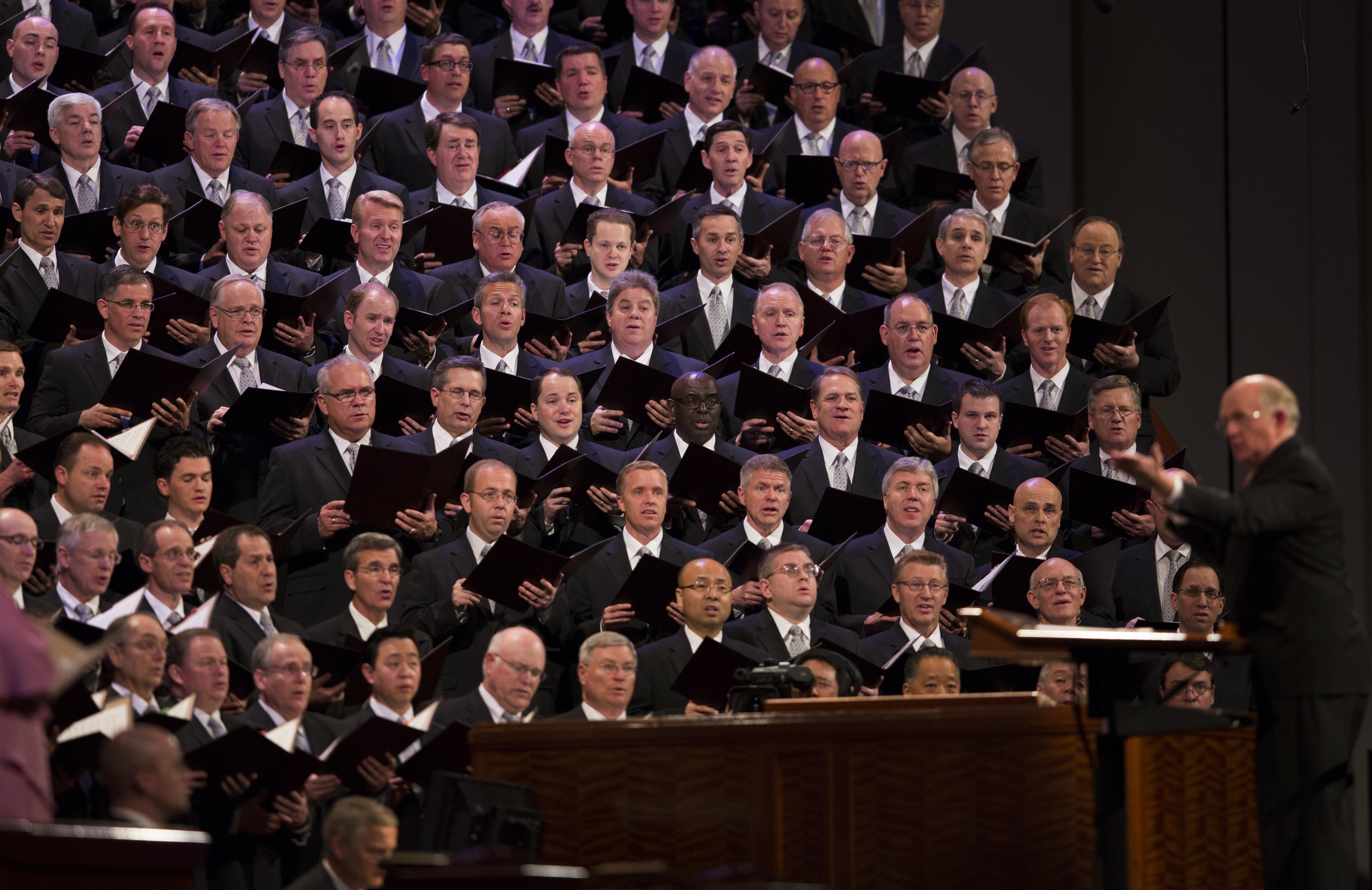 Rows of men from the Mormon Tabernacle Choir wearing dark suits and singing while Mack Wilberg conducts.